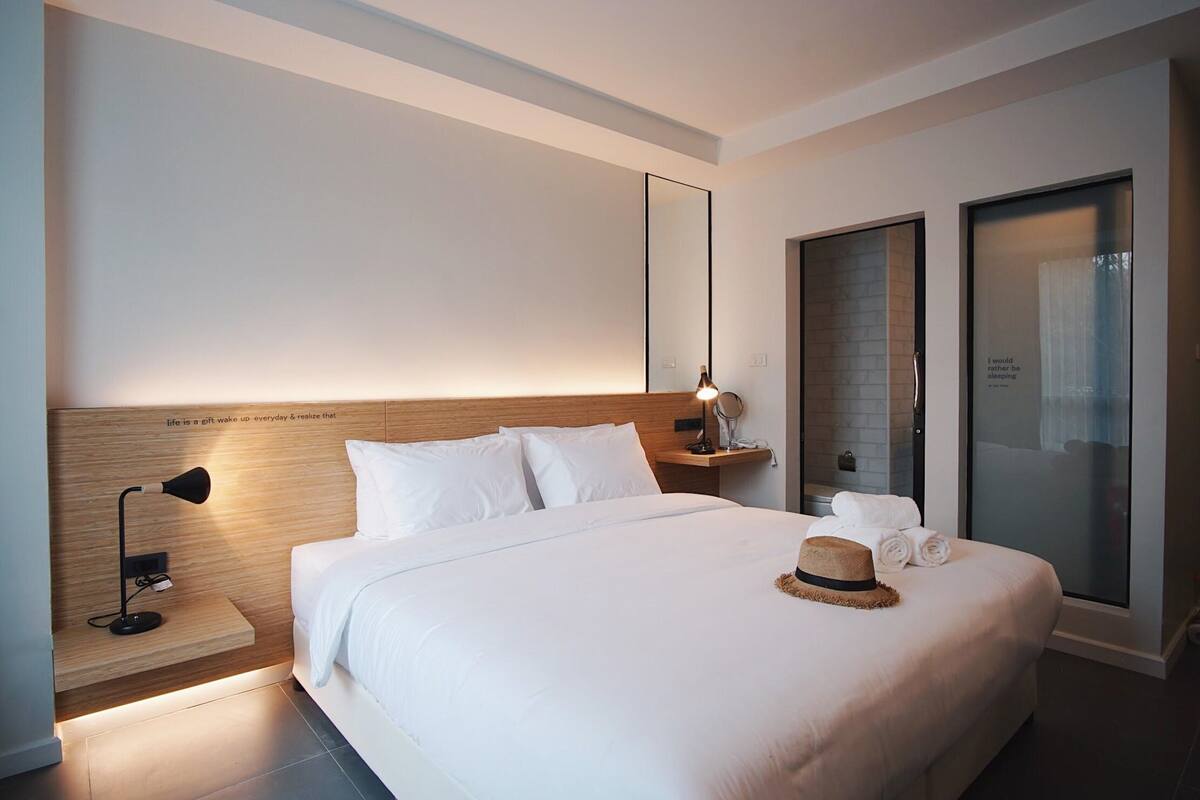 A modern hotel room with a large bed, a bedside table with a lamp, a mirror, and a view of the outside through a glass door.