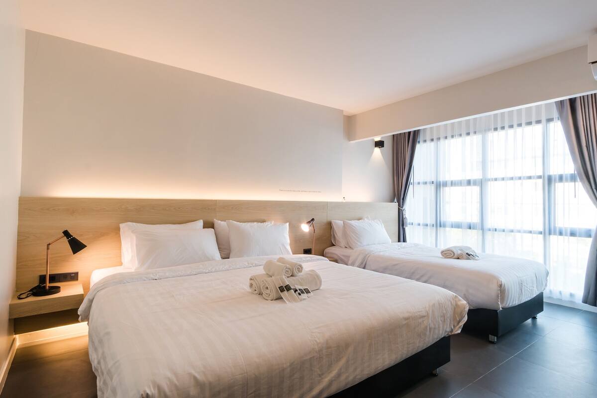 A hotel room with two beds, a bedside table with a lamp, and a large window with curtains.