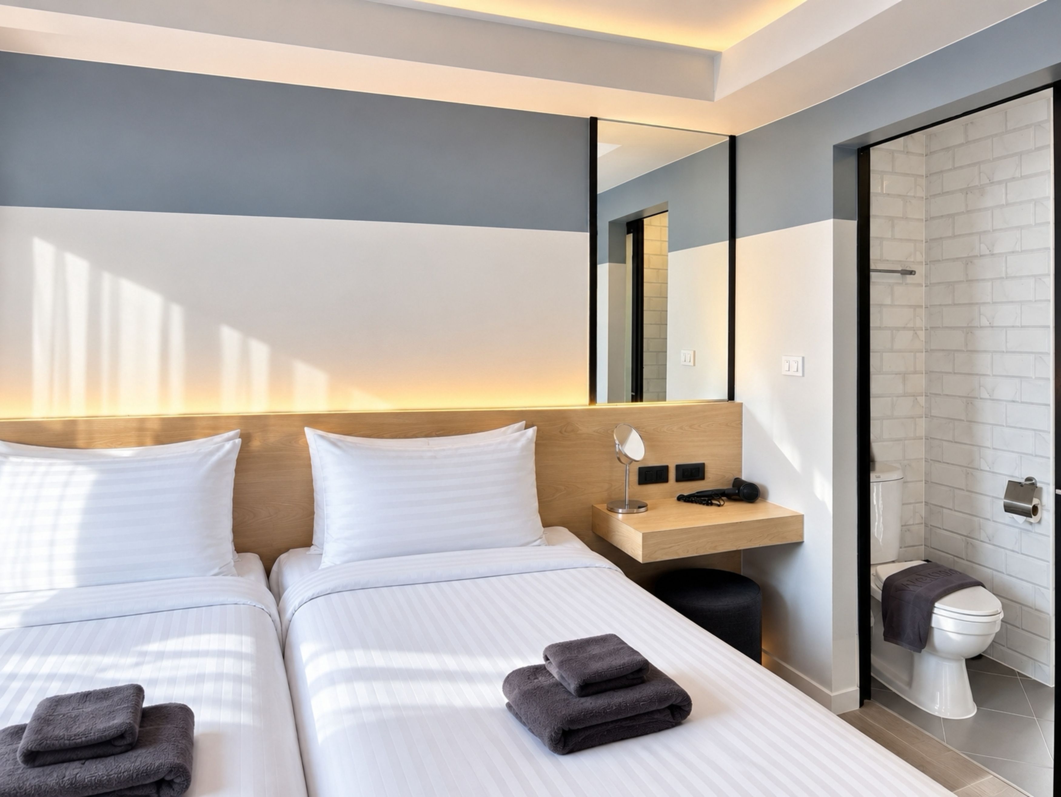A hotel room with two beds, a bedside table with a lamp, a mirror, a small shelf, and a bathroom with a toilet and shower.
