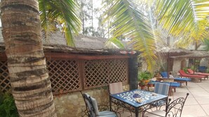 An outdoor patio with a table and chairs, surrounded by palm trees and a thatched-roof structure.