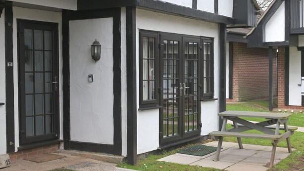 Un bâtiment à la façade blanche, aux ornements noirs et à la porte noire. On y trouve une fenêtre avec un grillage métallique et une petite table avec un banc.