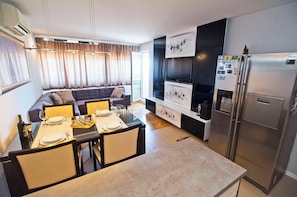 A modern kitchen with a built-in refrigerator, a dining area with a table set for two, and a living area with a sofa and curtains.