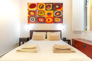 A neatly made bed with two pillows, a bedside table, a nightstand, and a red dresser in a hotel room.