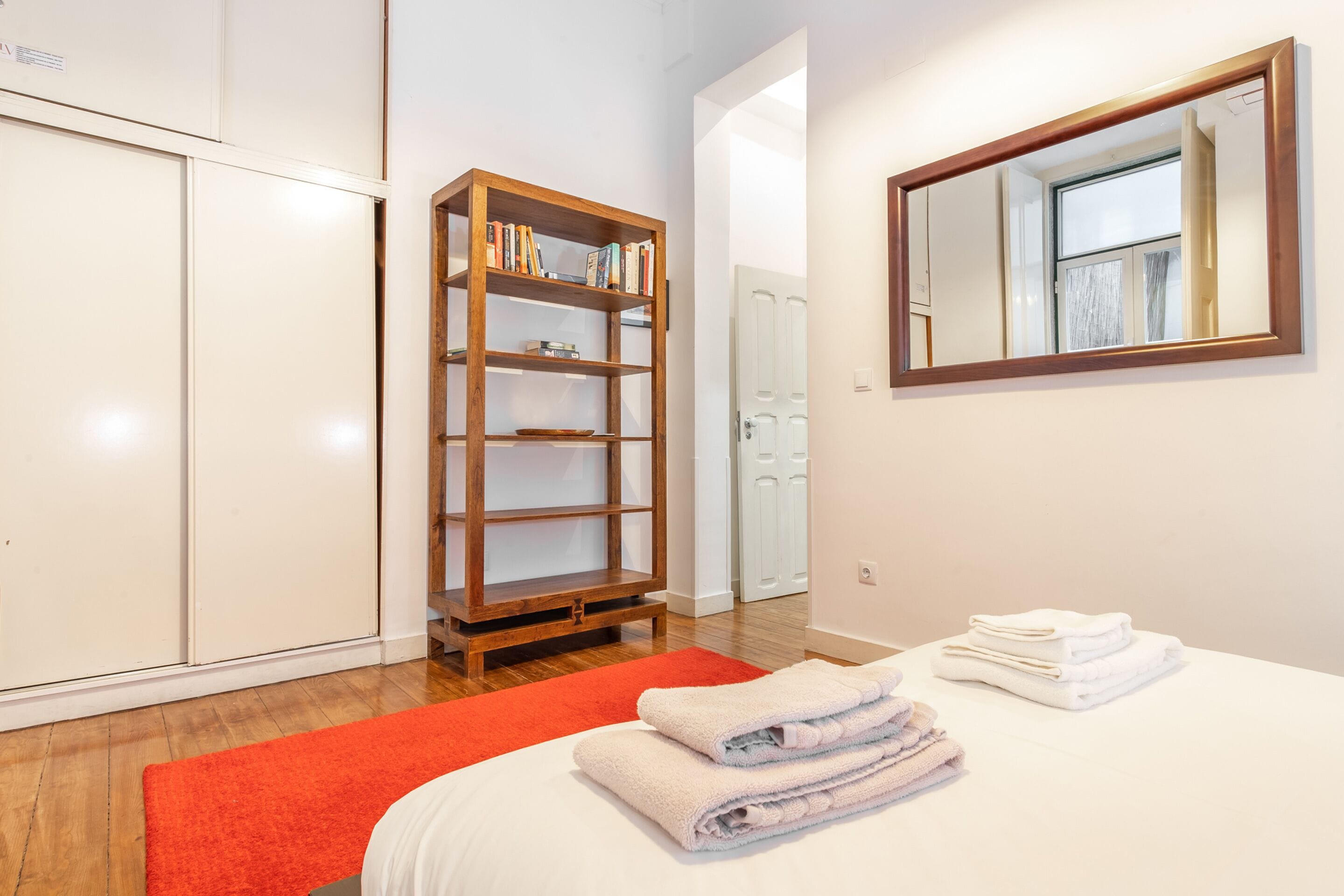 Een hotelkamer met een bed, een houten boekenkast, een spiegel en een rood tapijt.