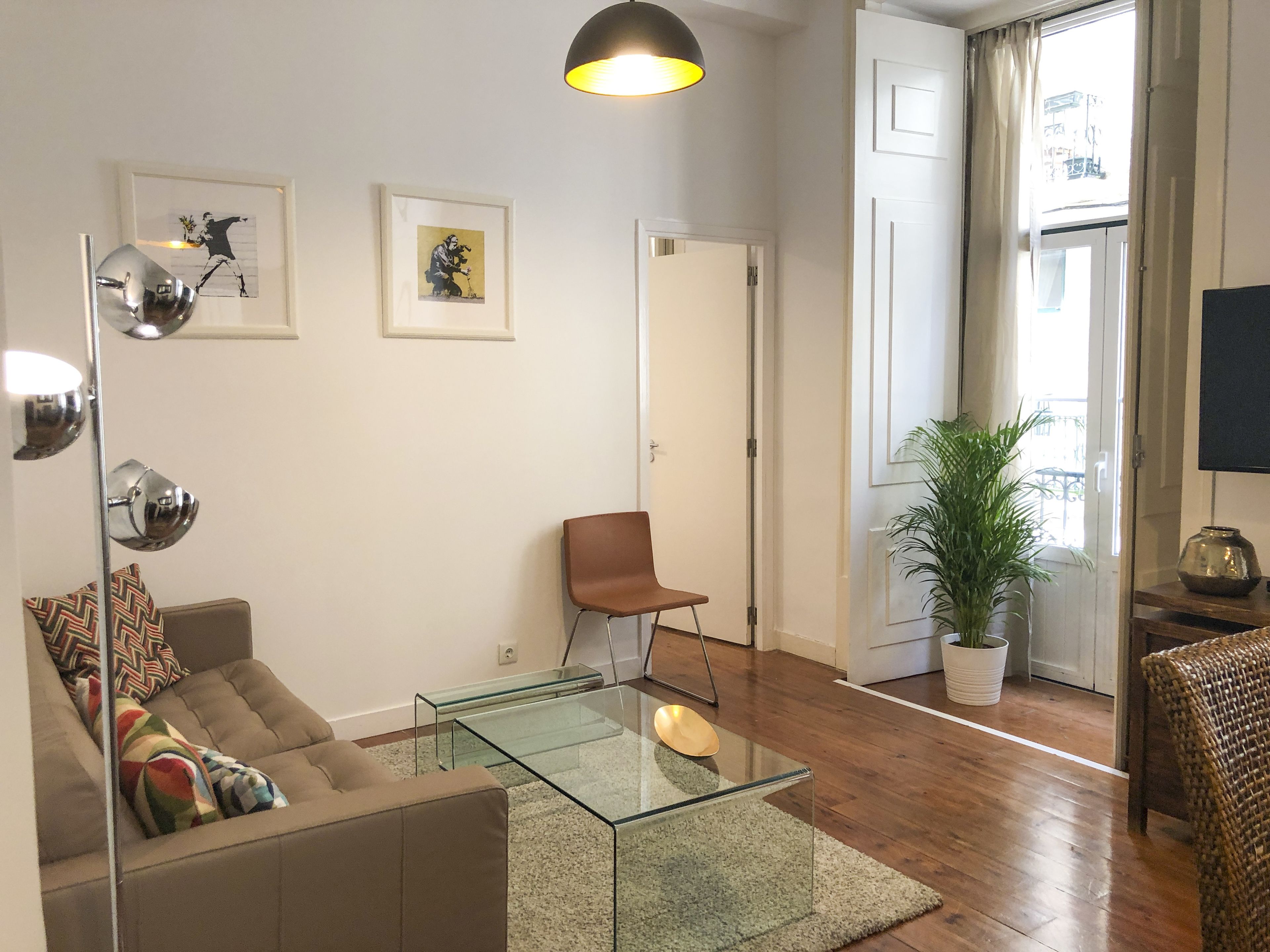Un salón moderno con una mesa de centro de vidrio, un sofá marrón, una silla y una lámpara de pie. Hay dos cuadros enmarcados en la pared y una planta en maceta cerca de la ventana.