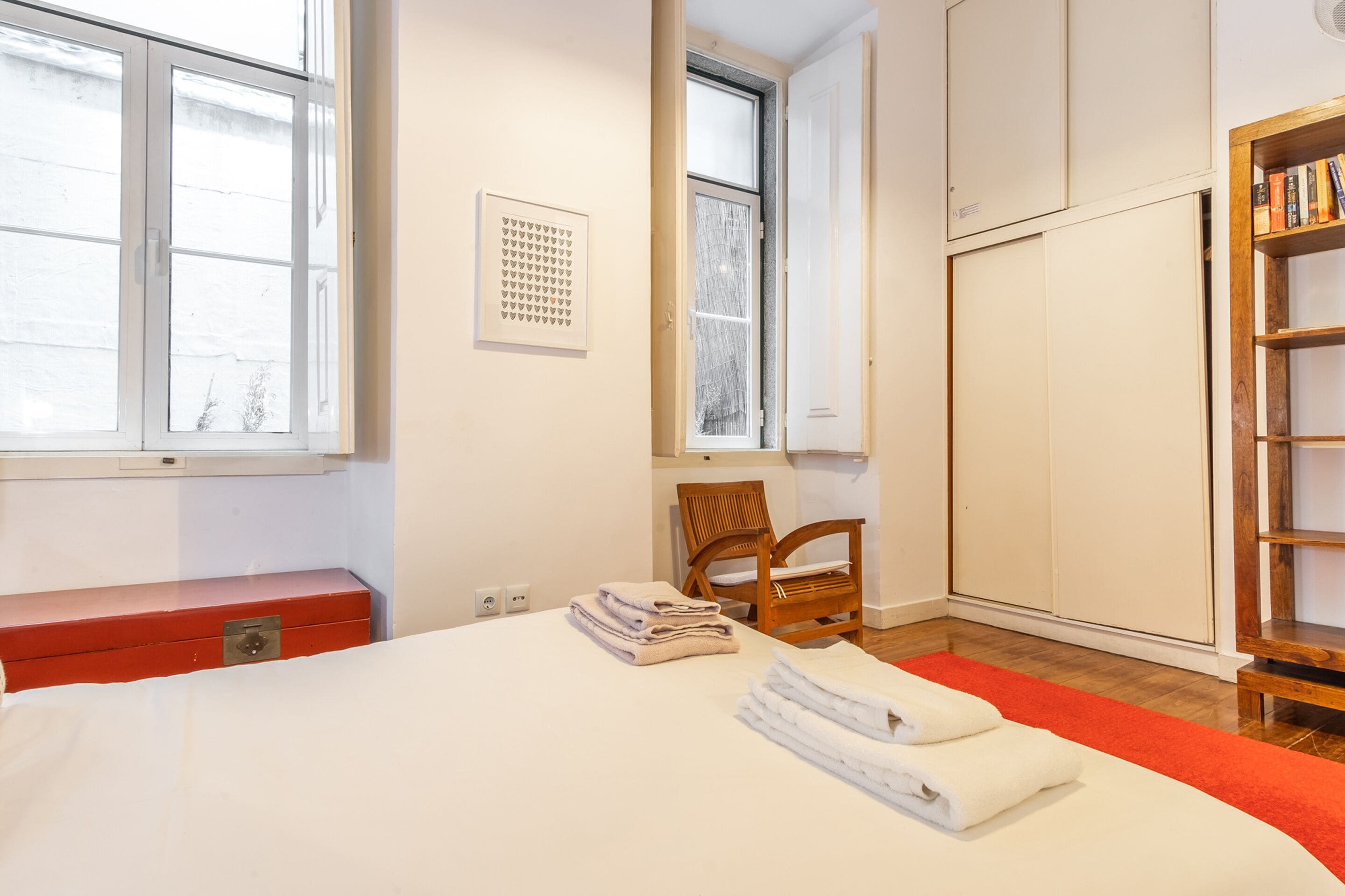 Una habitación de hotel con una cama, un aparador rojo, una silla de madera, una estantería y una ventana con vistas.