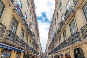 Una calle estrecha bordeada por edificios con balcones, un local visible con un letrero que dice "American Music Burger", y un cielo despejado arriba.
