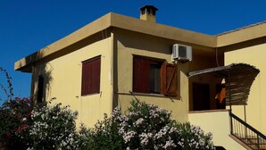 Il s’agit d’un bâtiment à deux étages, doté d’un toit plat, d’une cheminée et d’un appareil de climatisation. Il y a deux fenêtres avec des volets, dont l’une est ouverte. Le bâtiment est entouré de plantes en fleurs.