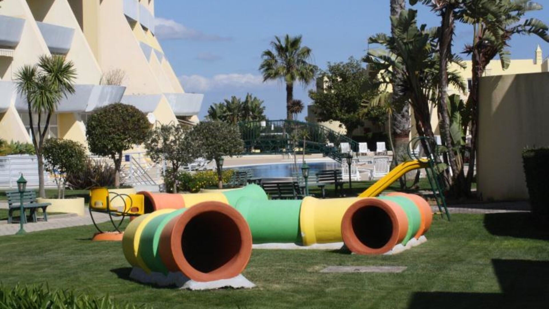 A playground with colorful tunnels and a slide, surrounded by palm trees and a pool.