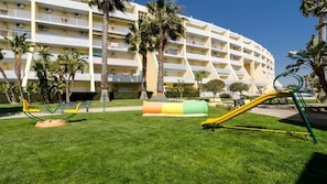 A hotel with a large, multi-story building, a grassy courtyard with playground equipment, and palm trees.