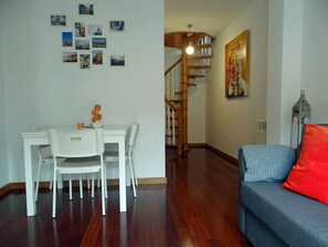 Uma área de refeições com uma mesa e cadeiras brancas, uma escada com corrimão de madeira e um sofá azul com uma almofada vermelha.