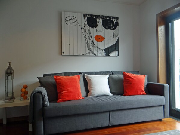 Uma sala de estar moderna com um sofá cinzento, almofadas vermelhas e brancas, um quadro abstrato na parede e uma mesa lateral com um vaso de vidro e laranjas.