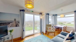 A room with a television, a sofa, a dining table, and a view of the sea.