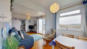 A living room with a sofa, TV, dining table, chairs, and a view through a large window. - D02 Greendune (D2), Riviere Towans (Hayle)