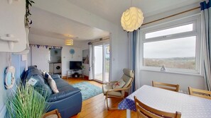 A living room with a sofa, TV, dining table, chairs, and a view through a large window. - D02 Greendune (D2), Riviere Towans (Hayle)