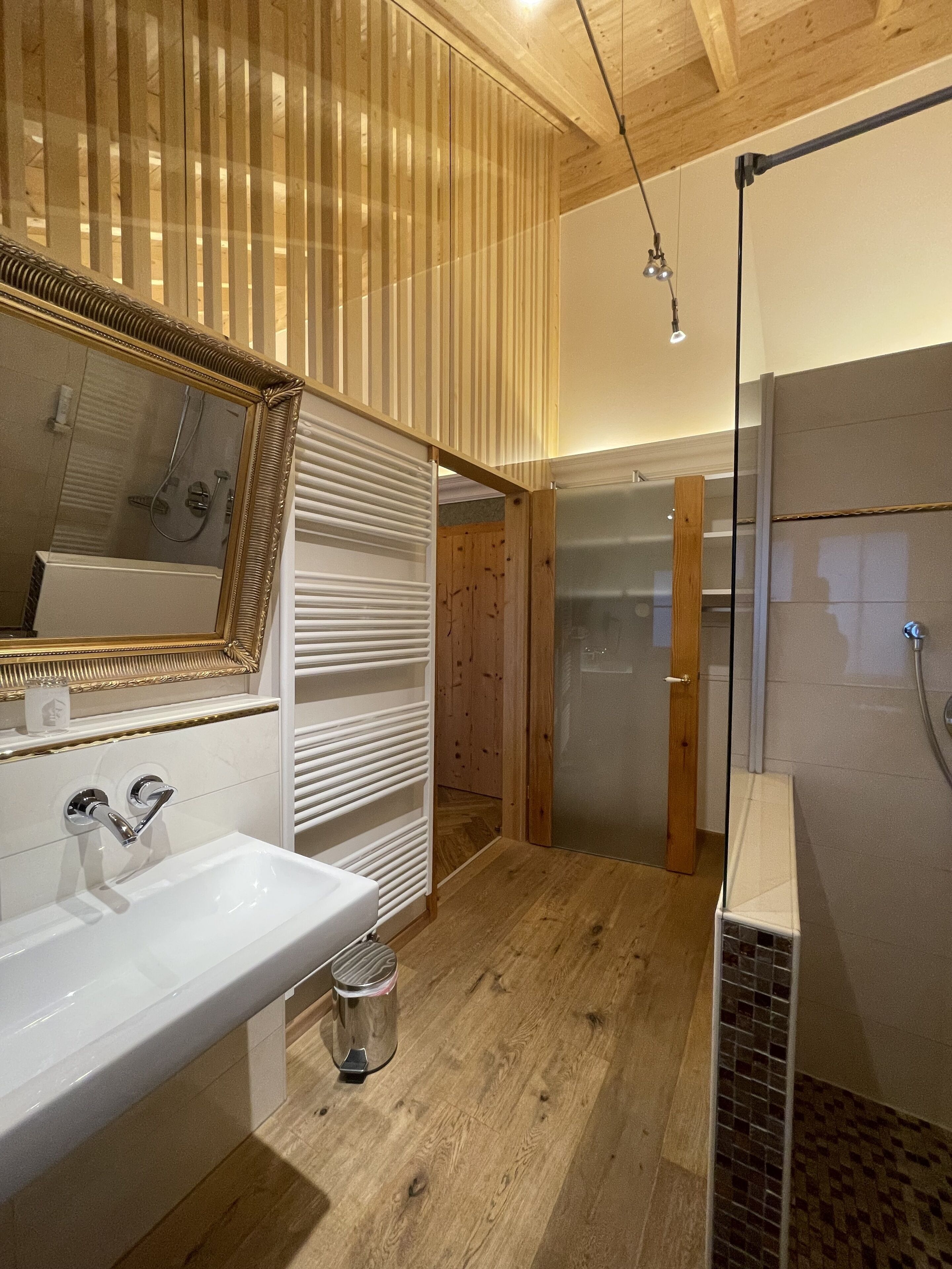 A modern bathroom with a wooden floor, a white sink, a large mirror, and a glass shower enclosure.
