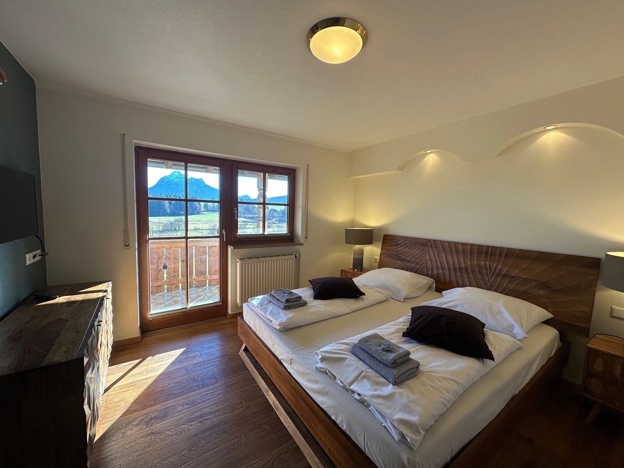 Una camera da letto con due letti, una testiera in legno, una finestra vista montagne, un termosifone e un comodino con una lampada.