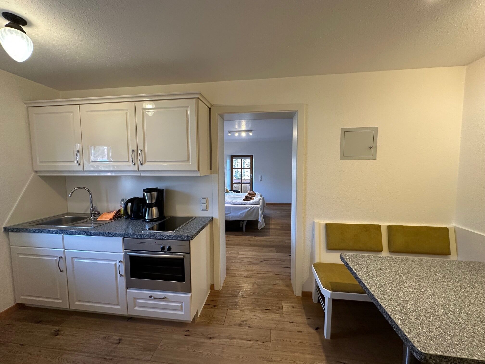 A compact kitchen with a sink, stove, and countertop, leading to a bedroom with a bed and a window.