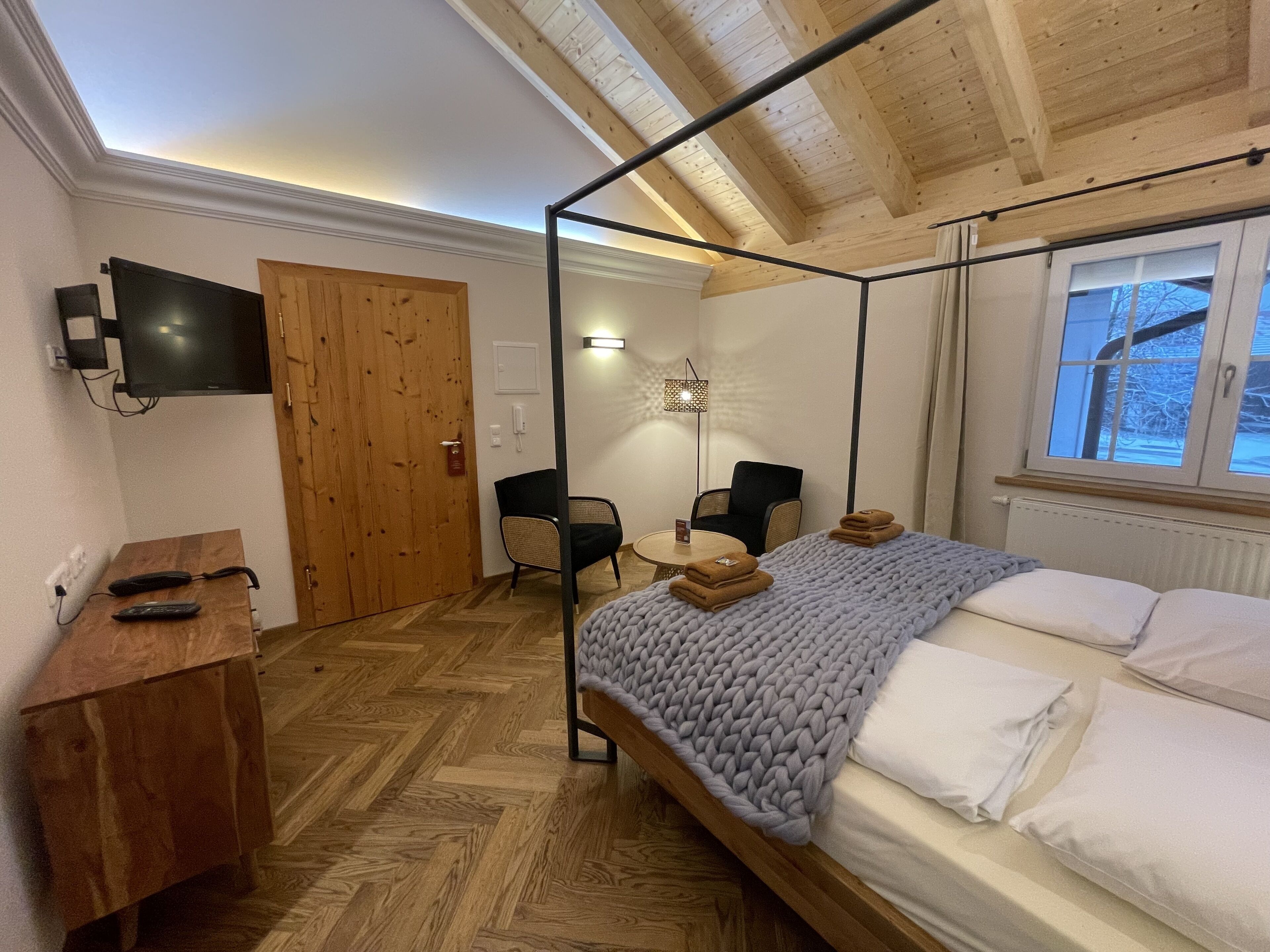 A bedroom with a four-poster bed, a desk with a chair, a television, and a window with a view of the outside.