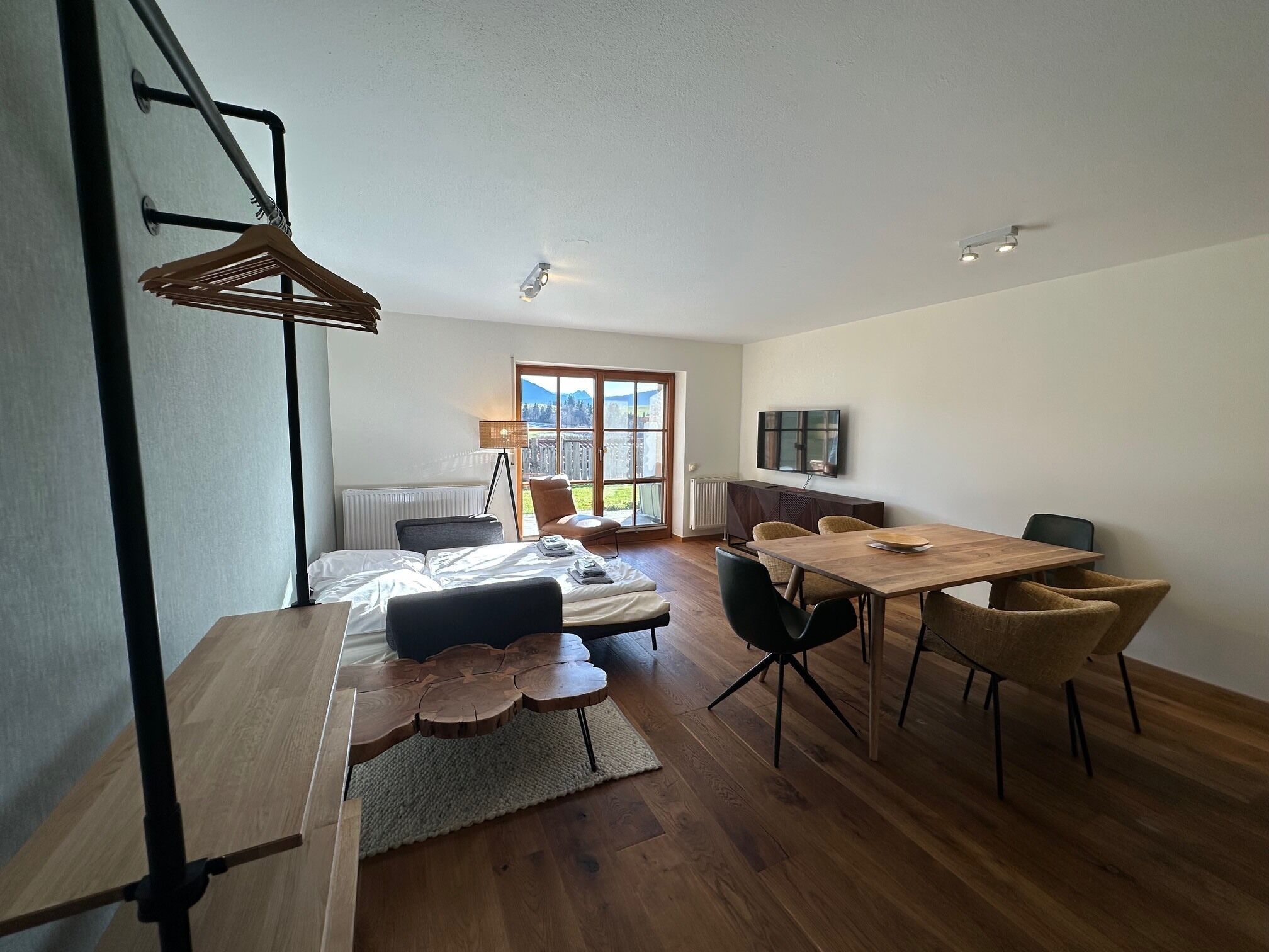 Een moderne, goed verlichte kamer met een houten eettafel, stoelen, een bed en een televisie.