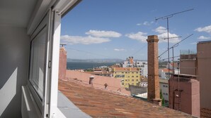Une vue depuis une fenêtre donnant sur un paysage urbain, avec des bâtiments, un ciel dégagé et une étendue d’eau au loin.