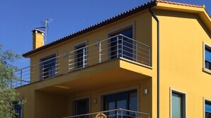 Un edificio de dos pisos con fachada amarilla, un balcón con mobiliario de mimbre y una escalera que lleva a la entrada.