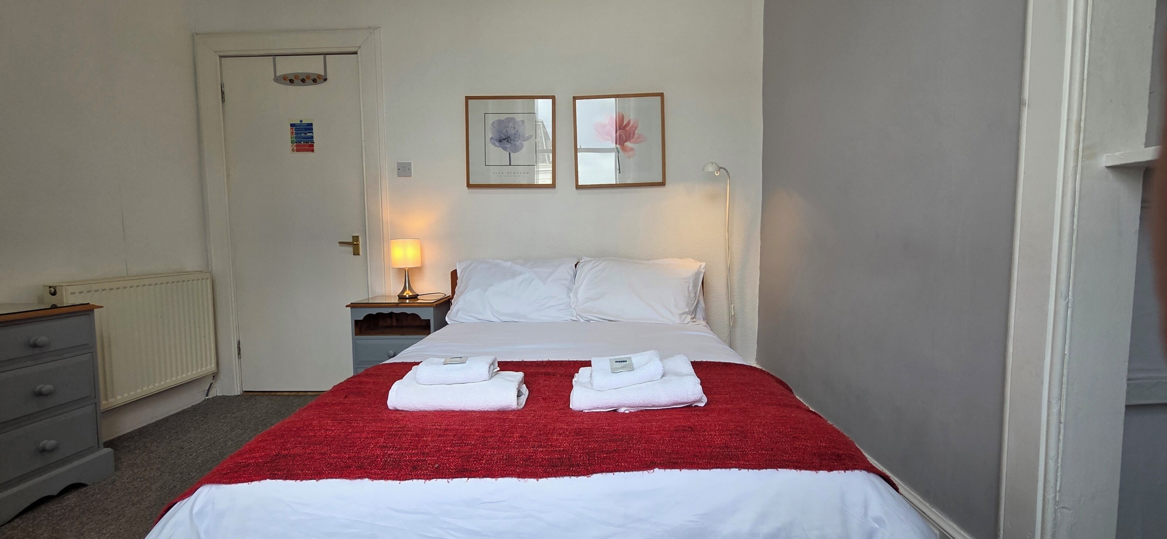 Une chambre d’hôtel avec un lit, une table de chevet, une lampe, une porte et deux tableaux encadrés au mur.