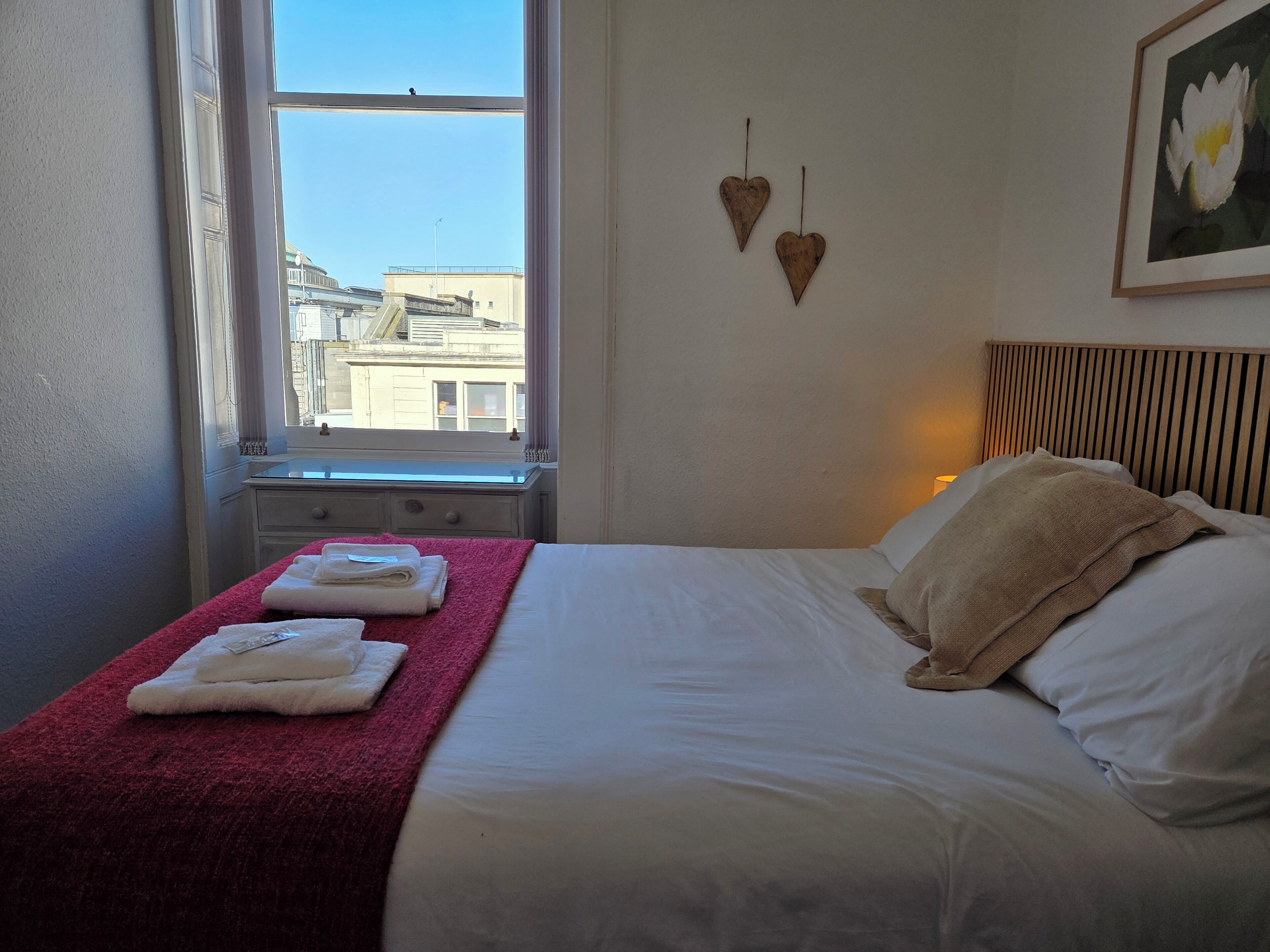 A hotel room with a bed, a window, a red towel, and a wall-mounted picture.