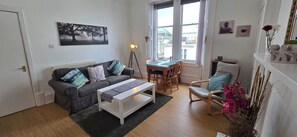 Een woonkamer met een bank, een salontafel, een eettafel met stoelen, een lamp, een raam met uitzicht en een plank met decoratieve objecten.