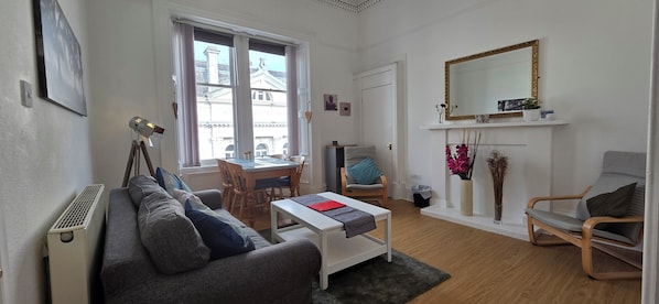 A compact living space with a sofa, a dining table, a chair, a small coffee table, a radiator, a window with a view of buildings, and a mirror on the wall.