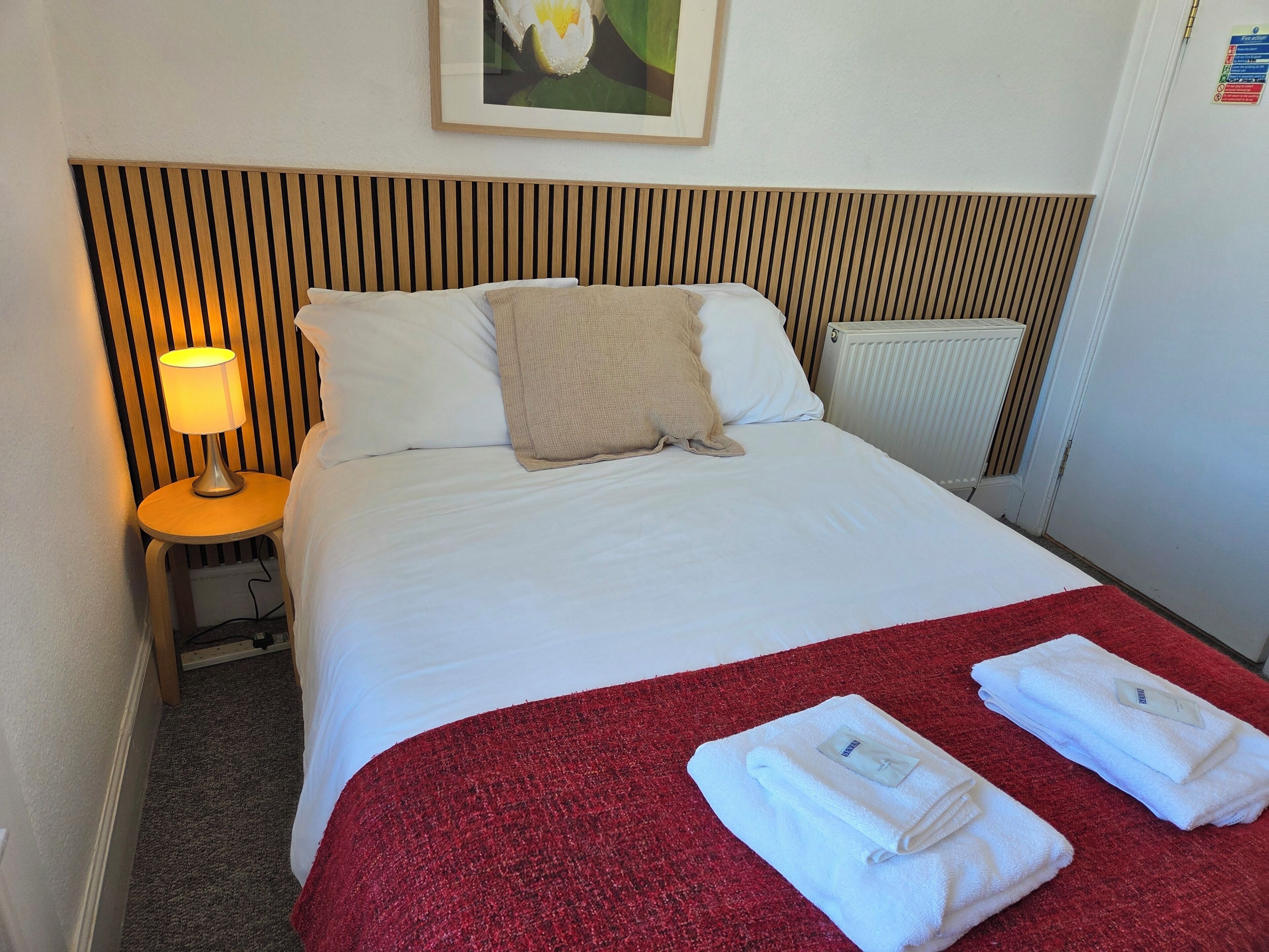 Habitación de hotel con cama, una manta roja, dos toallas dobladas, mesita de noche con lámpara y radiador.