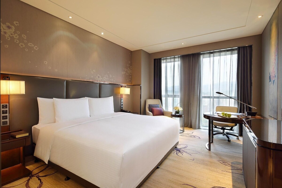 Quarto de hotel com uma cama grande, uma escrivaninha e uma cadeira. Há uma janela com cortinas e vista para a cidade.