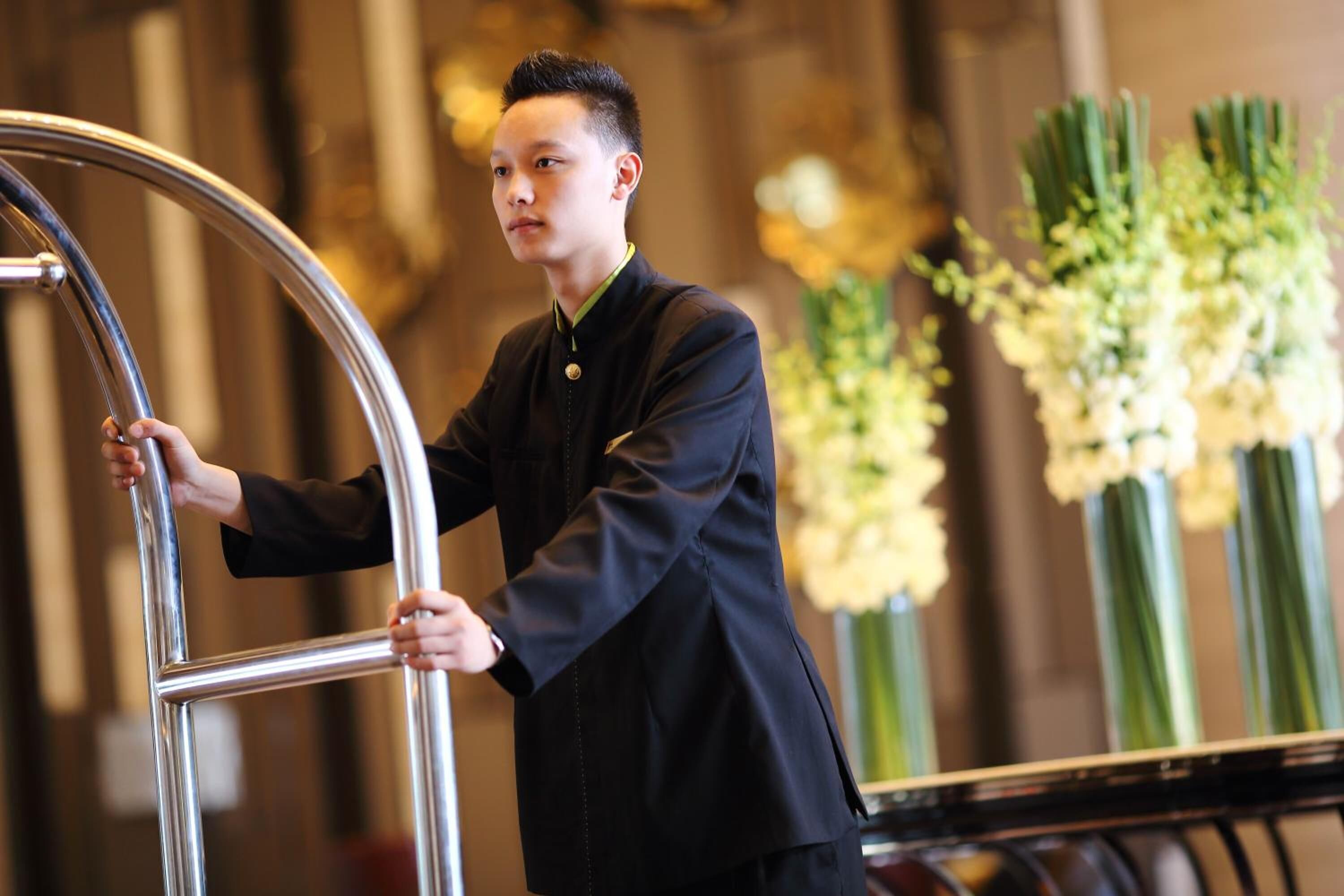 Een persoon in een zwart uniform staat naast een metalen leuning, waarschijnlijk in een hotellobby met bloemversiering op de achtergrond.