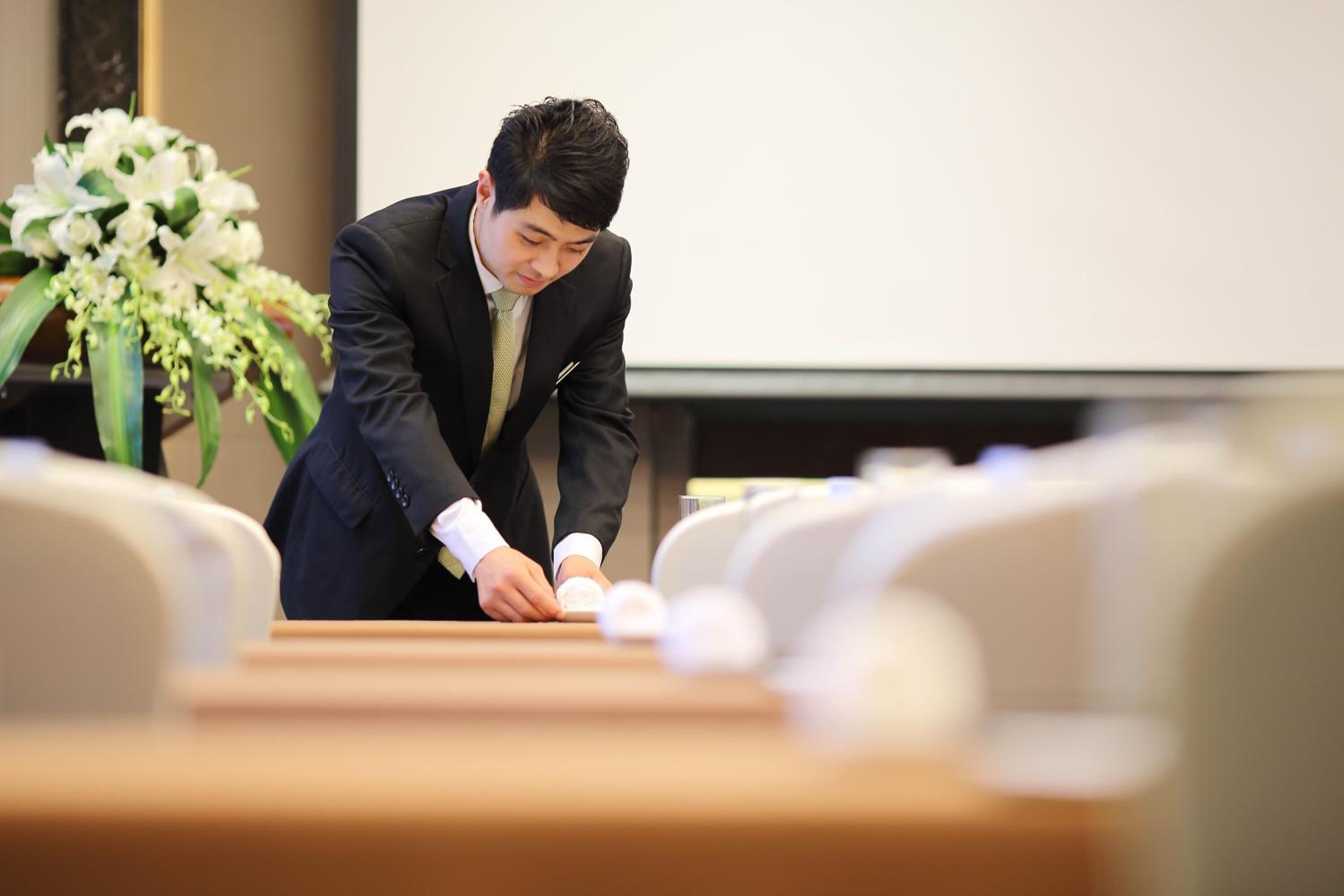 A man in a suit sitting at a table with a laptop, in a room with chairs and a whiteboard in the background.