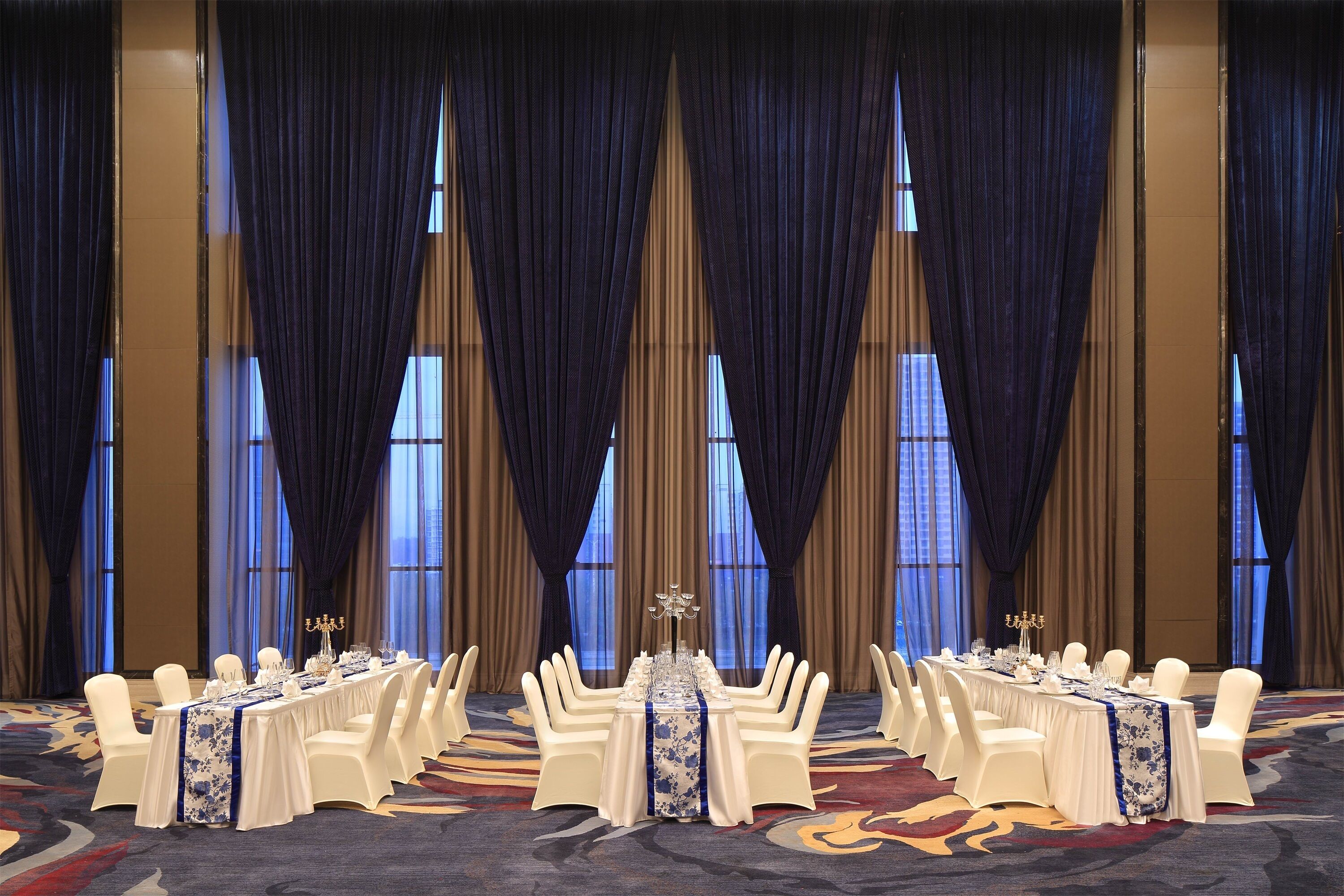 A banquet hall with a long table set for a formal event, flanked by tall curtains and large windows.