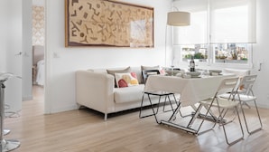 A modern living room with a white sofa, a dining table with chairs, a large abstract painting, and a city view through large windows. - Smart Cuzco Castellana Penthouse (Madrid)