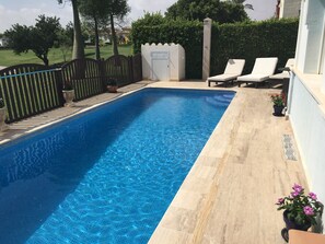 A rectangular swimming pool with clear blue water, surrounded by a tiled deck. There are lounge chairs and potted plants along the poolside.