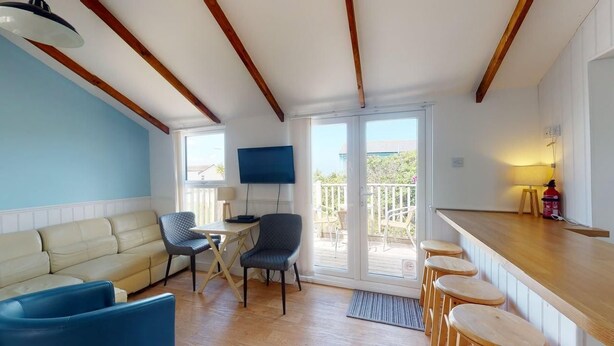 A room with a wooden ceiling, a sofa, a chair, a table, a TV, a balcony with a railing, and a bar area with stools.