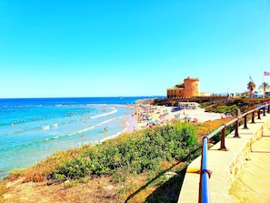 Une plage avec un bâtiment qui ressemble à un château, une promenade et une eau d’un bleu limpide.