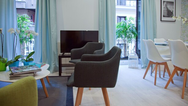 Uma sala de estar moderna com uma TV de tela plana, duas poltronas cinza escuro, uma mesa de centro branca e uma área de jantar com cadeiras brancas.