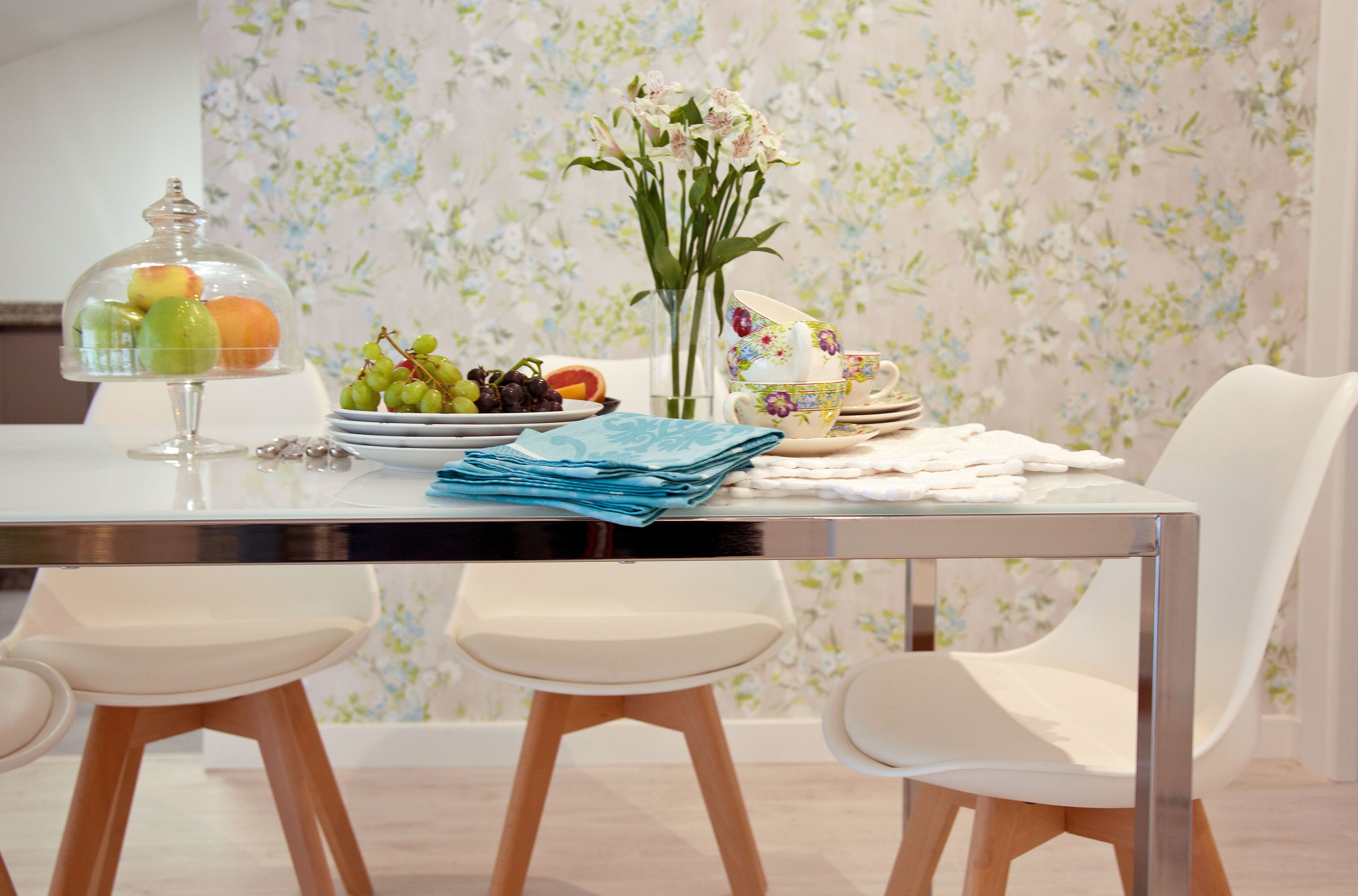 A dining table with a floral wallpaper background, a glass fruit display, and white chairs.