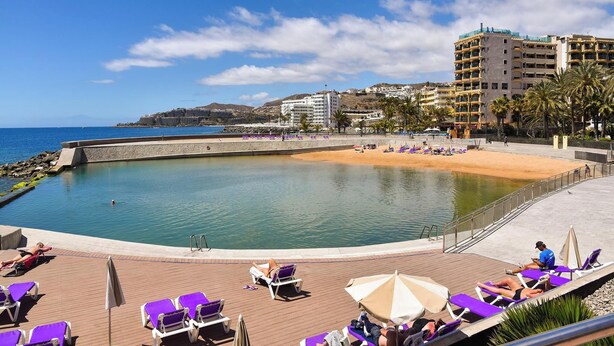 Ein Poolbereich mit Liegestühlen und Sonnenschirmen, im Hintergrund ein Strand, und Gebäude umgeben die Szene.