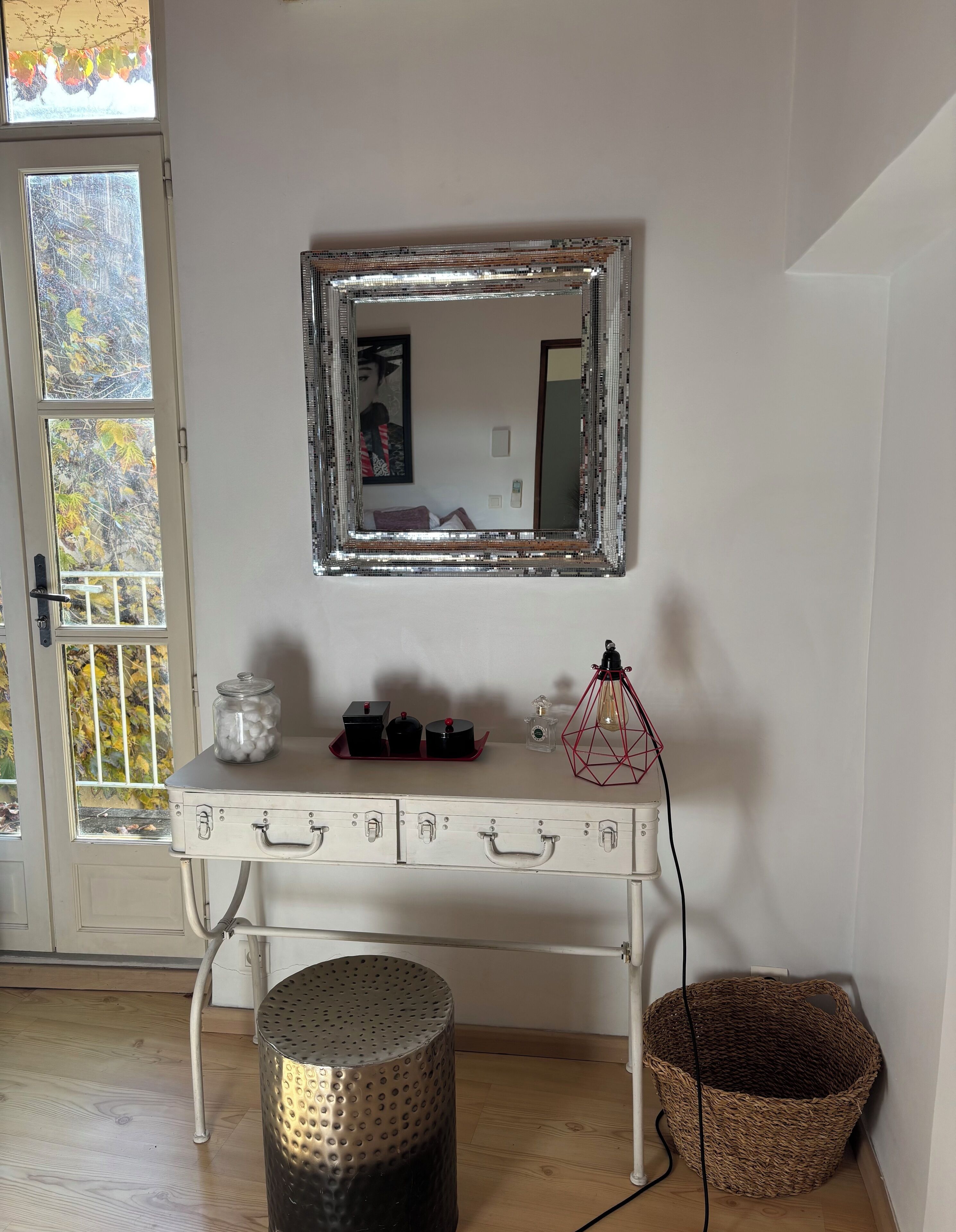 A white dresser with drawers, a mirror with a metallic frame, a small lamp, and a basket on the floor.