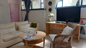 Una sala de estar con un sofá, una mesa redonda para café, un sillón de mimbre, un escritorio con televisor y una puerta rosa.