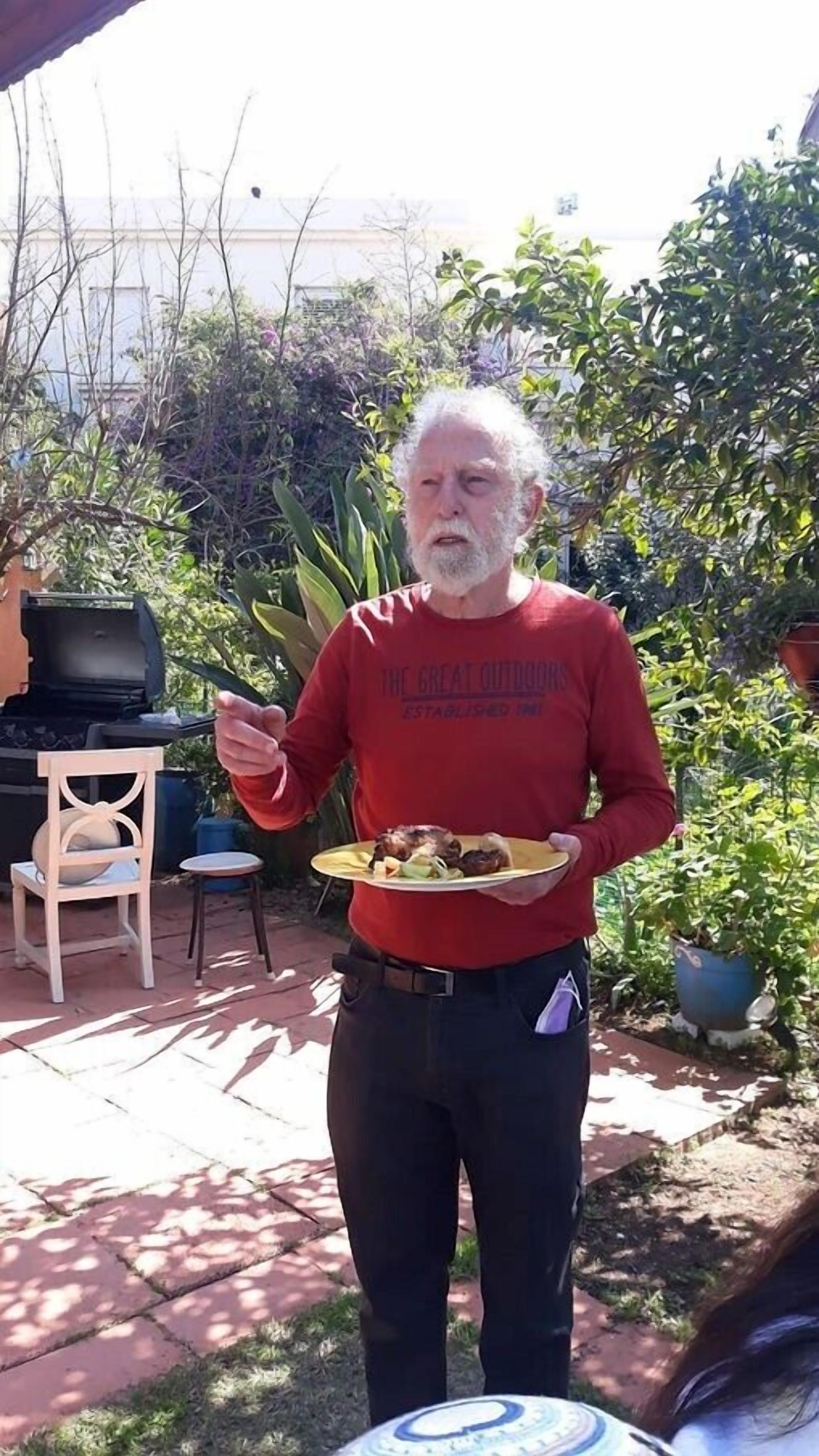 A person standing outdoors holding a plate of food, with a grill and chairs in the background.
