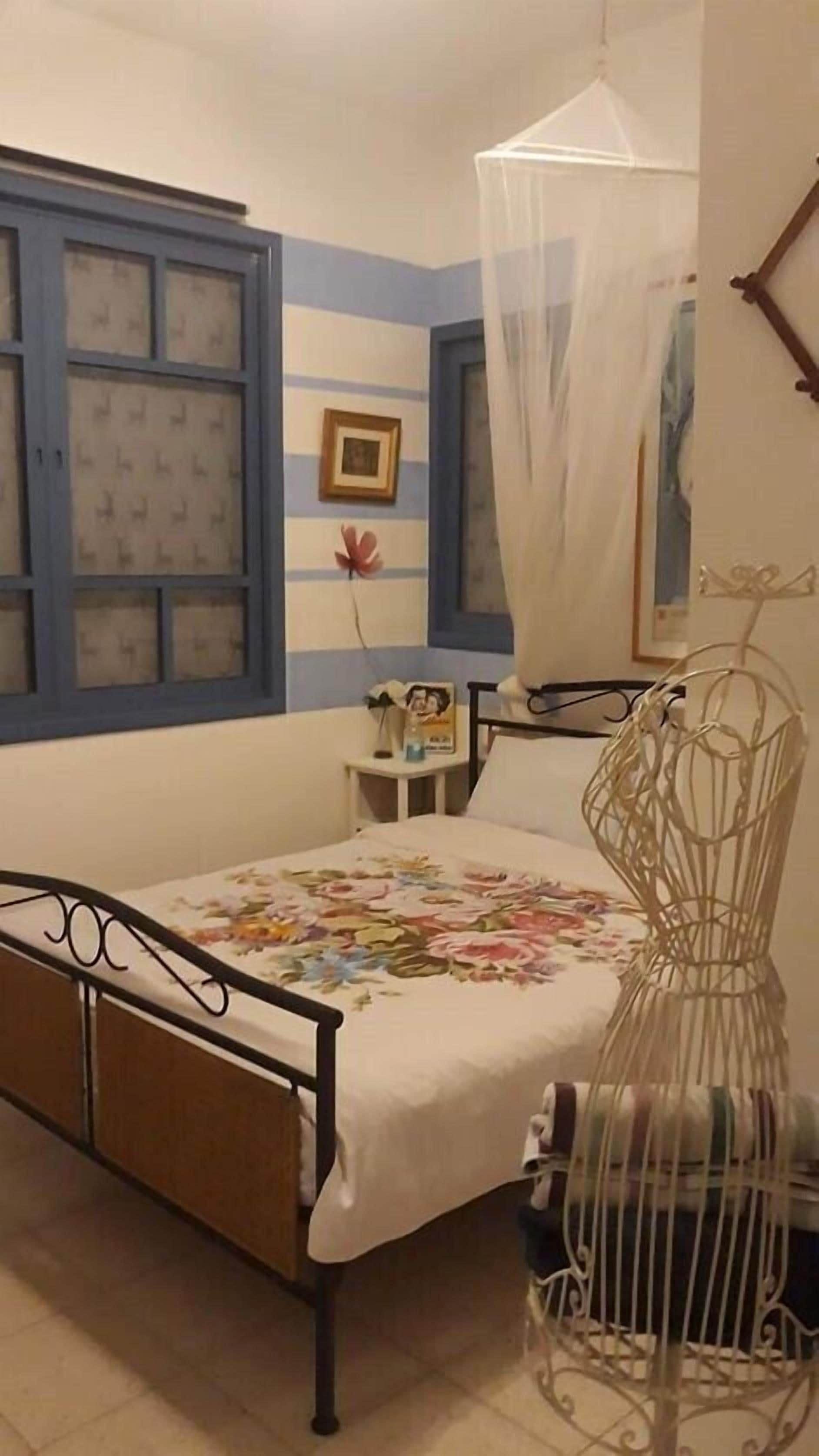 A bedroom with a bed, a bedside table, a nightstand, a window with blue shutters, a framed picture on the wall, and a decorative birdcage.