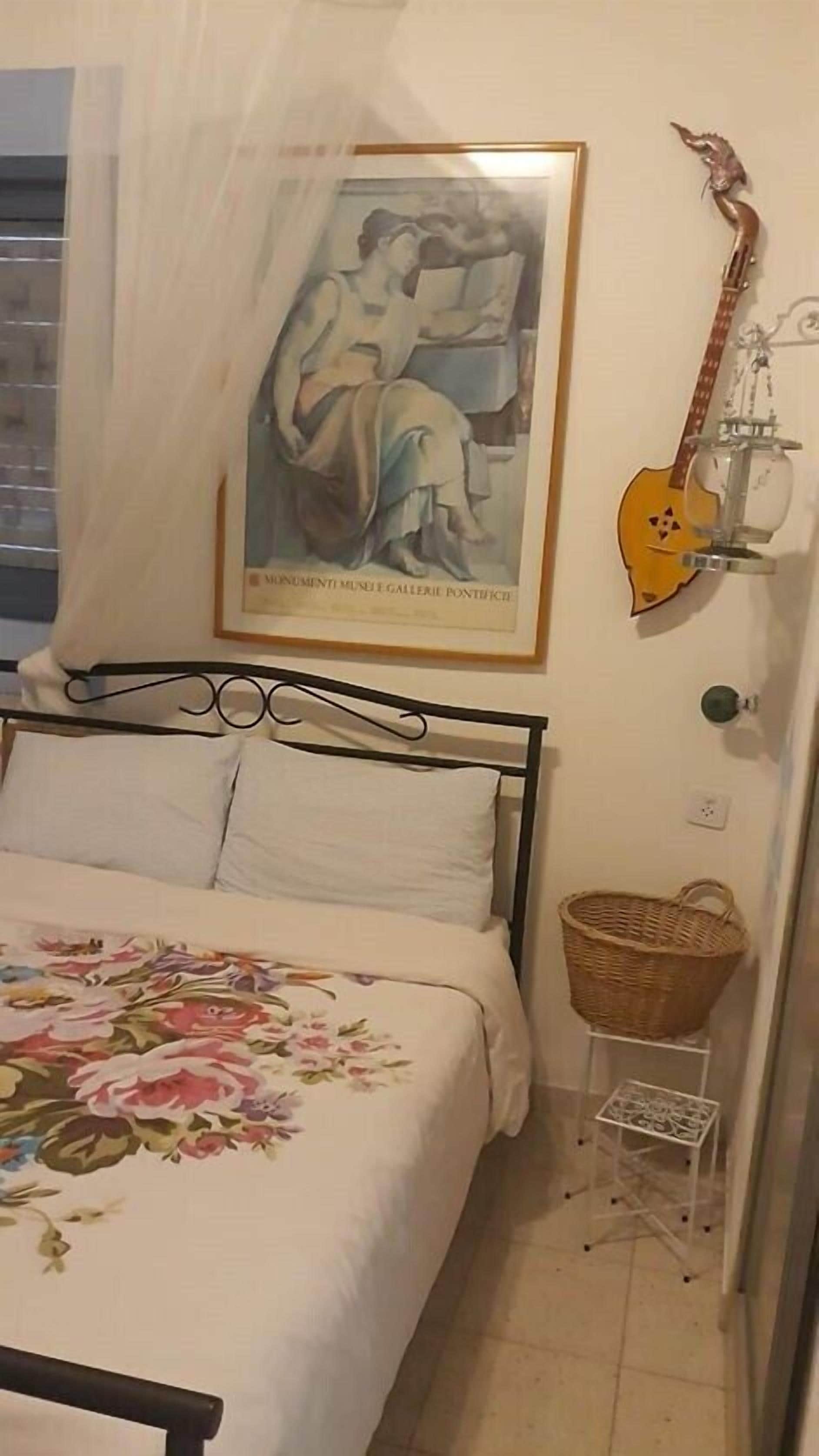 A bedroom with a bed, a framed painting, a guitar, and a wicker basket.