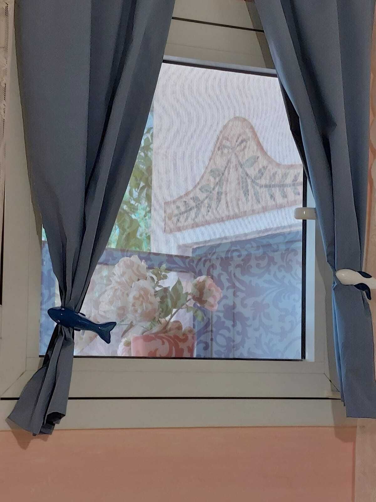 A window with grey curtains tied back, revealing a view of a balcony with a patterned railing and a vase of flowers.