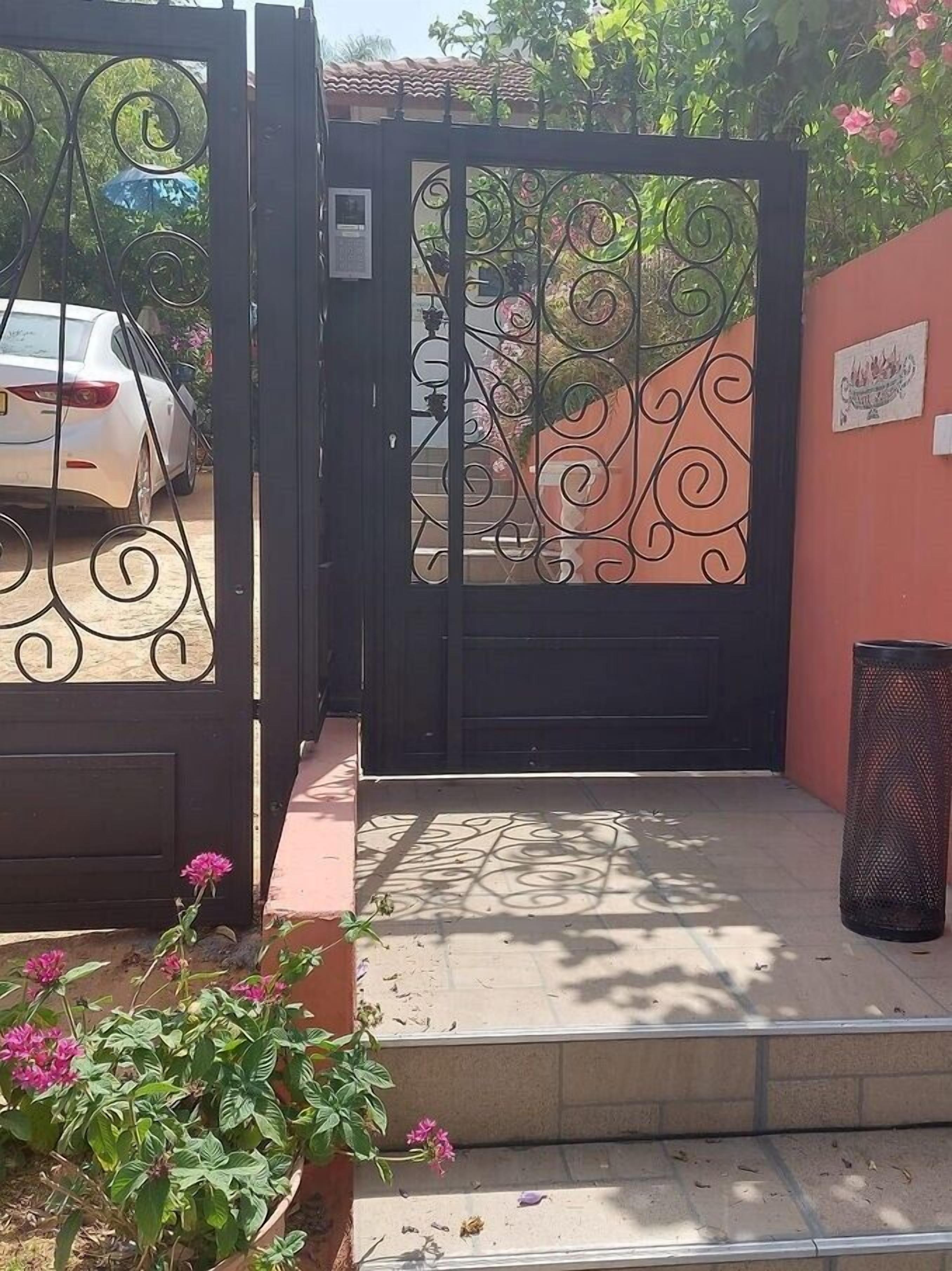 Uma entrada fechada por grades com um portão de metal decorativo, um carro branco estacionado na frente e uma lixeira à direita.