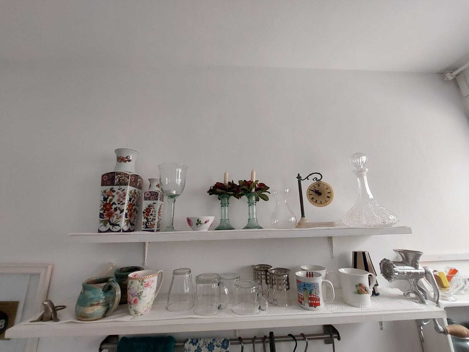 Een keukenkastje met diverse glazen, waaronder een vaas met bloemendesign, een heldere glazen kan en een klok.